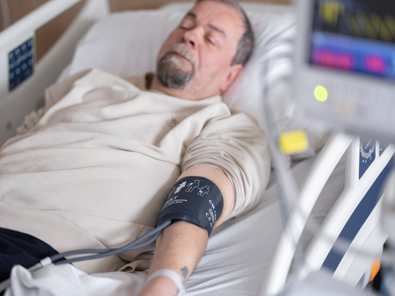 Elderly man resting in hospital bed with blood pressure monitor attached, symbolizing healthcare and medical care.