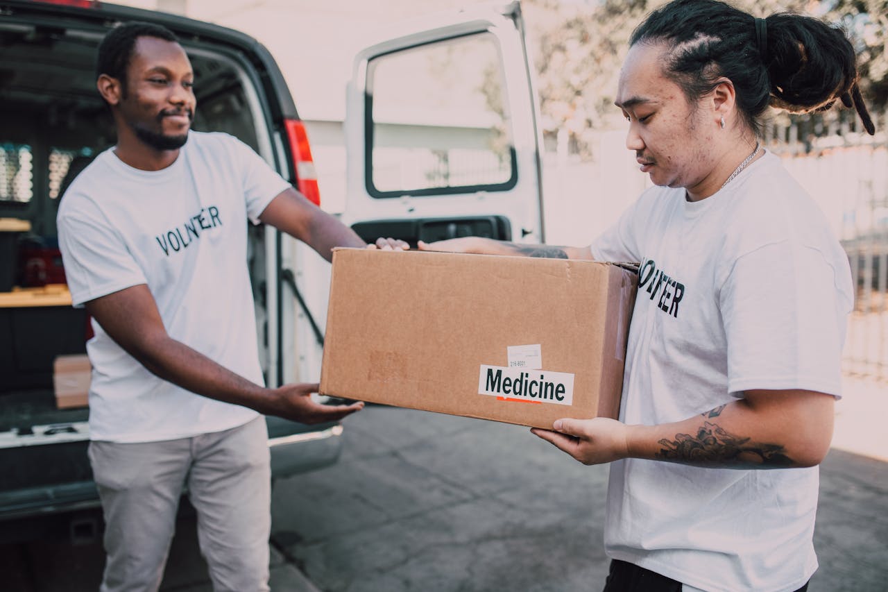 Two volunteers distribute a box of medicine from a van, emphasizing charity and teamwork.