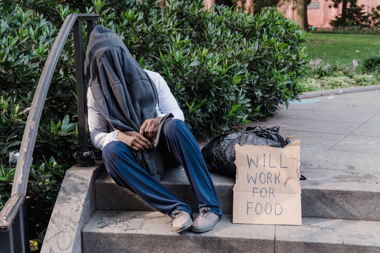 A homeless person sitting on outdoor stairs, covered by a cloth, with a sign reading 'Will Work for Food'.
